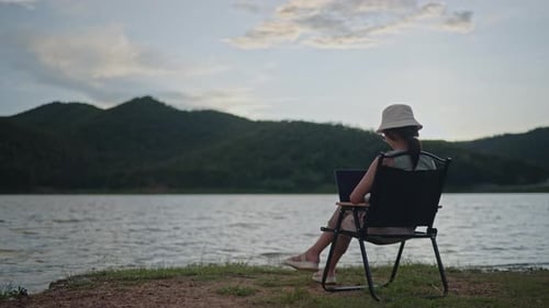 Mulher trabalhando no laptop à beira do lago e da montanha à noite com pôr do sol dourado