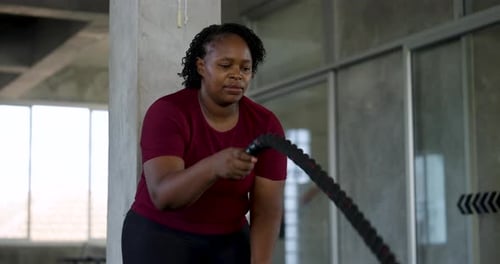 Front view of African American plus size woman making battle rope waves in modern gym. Curvy female