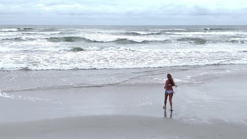 Woman Walking on Sandy Beach with Ocean Surf and Cloudy Sky Drone Video