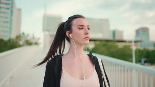 Young woman in wired headphones listens to music and walks along the bridge