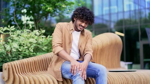 Young man suffers from painful severe knee joint ache while sitting on a bench near an office