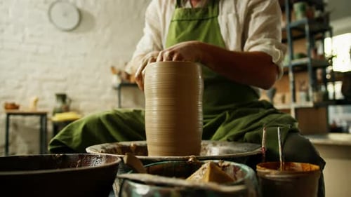 Close-up of potter's hands covered with clay making beautiful vase on throwing wheel in pottery