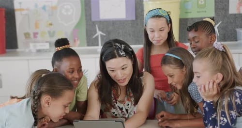 Happy diverse female teacher with schoolgirls using tablet in classroom at elementary school