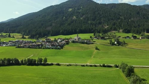 An alpine village lies nestled at the base of a forested mountain, with a prominent church and steep