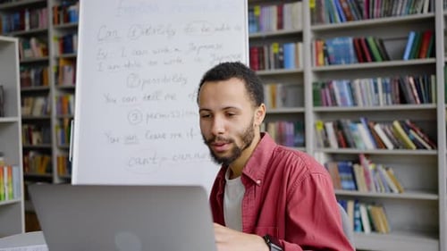 Multiethnischer Lehrer sitzt mit Laptop am Tisch und erklärt während des Online-Unterrichts eine Lektion auf Flipchart