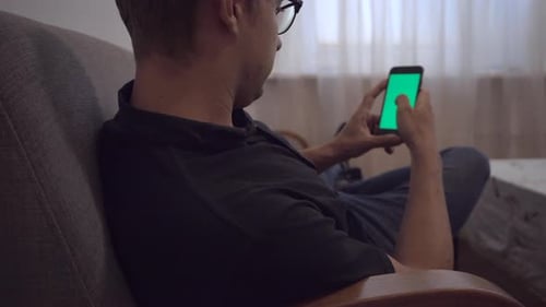 View on young man sitting on couch with green screen smartphone