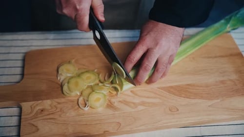 Close-up of hands slicing leek on wooden board with fresh ingredients around