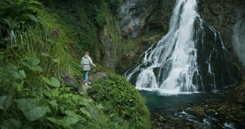Woman traveler hiking to a waterfall in the highlands, raises her hands up, enjoys nature and life.