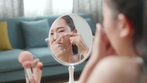 Reflection In A Mirror Worried Young Asian Woman Popping Pimples While Applying Makeup At Home