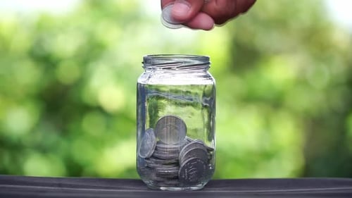 Hands putting coins in a glass container with a blurred green nature background. Saving concept. Eco