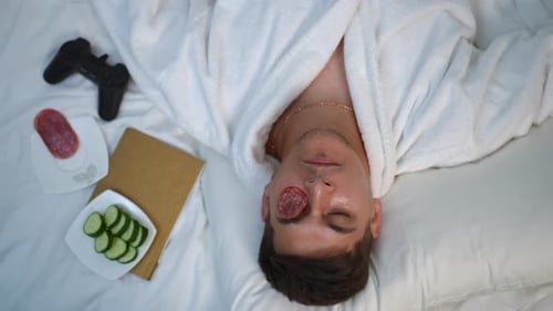 Top View A Funny Young Guy in a White Bathrobe Eats While Lying on the Bed in the Bedroom at Home