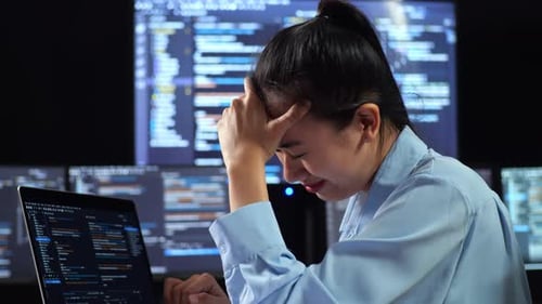 Programadora asiática que tiene dolor de cabeza mientras escribe código con una computadora portátil con varios monitores