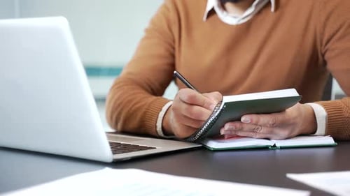 Primer plano de manos masculinas tomando notas en el cuaderno mientras usan el portátil en el escritorio de trabajo en el lugar de trabajo