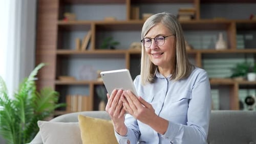 Senior gray haired female watches a video stream on a digital tablet while sitting on the sofa