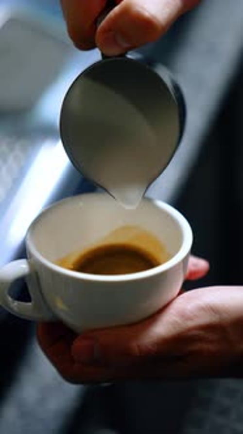 Male hands hold a white coffee cup filling it with milk. Barista preparing latte drink. Close up.