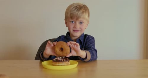 A Young Boy Holds a Chocolate Donut in His Hands While Another One Lies on a Plate in Front of Him