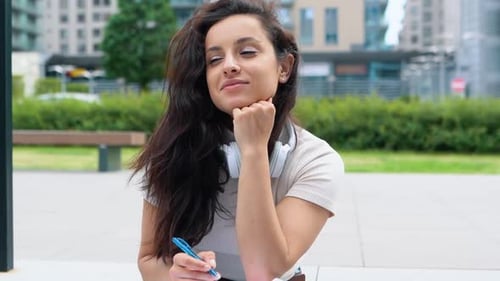 Happy Thoughtful Female Student Sitting in City and Writing in Copybook