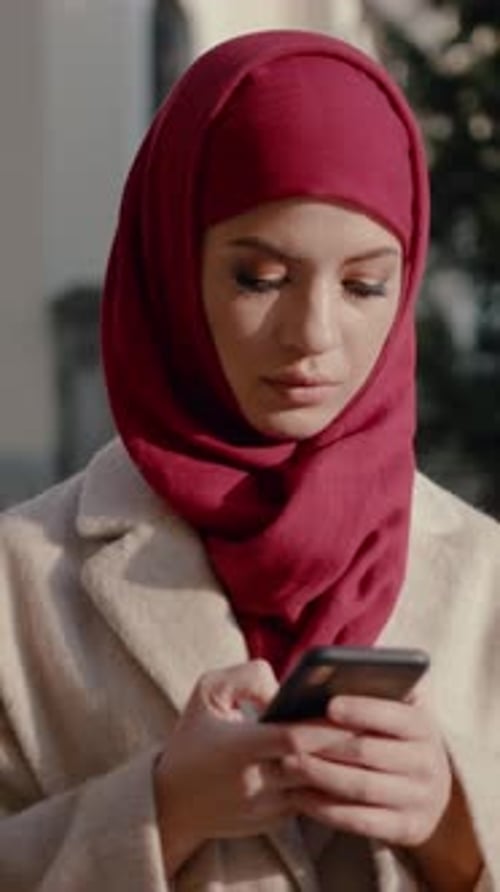 A Young Woman Wearing a Red Hijab is Seen Casually Texting on Her Smartphone Outdoors
