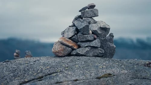 Close-up shot of the stone cairn. Gloomy landscape in the background. Grey clouds above. Slow-motion
