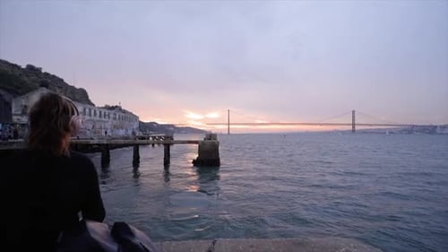 Jovem mulher sentada à beira do rio Tejo, olhando para a água e ouvindo música.
Lisboa