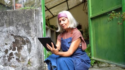 Woman Sitting by Greenhouse Door Using a Tablet