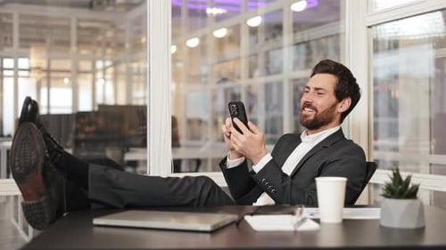 Happy Businessman Relaxing with Feet Up on Desk Using Phone in Office