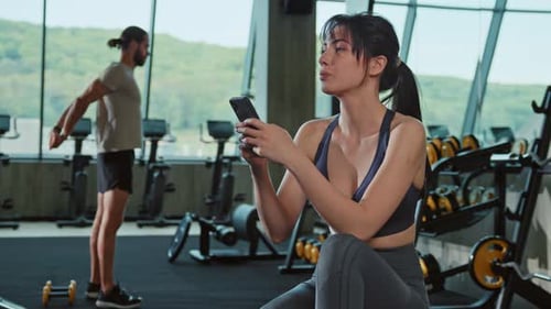 Shooting of Sporty Young Smiling Woman in Gym Sitting on Bench Press Using Smartphone for Chatting