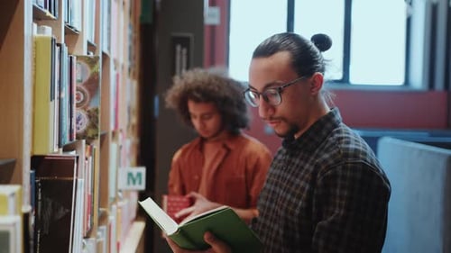 Male Student Taking Book from Shelf and Reading It in Library