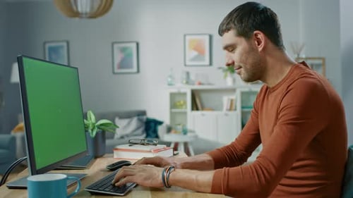 Handsome Man Sitting at His Desk at Home Uses Personal Computer with Mock-up Chroma Key Screen.
