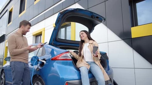 Happy young couple smiling and talking while drinking coffee standing outdoor at EV charging station