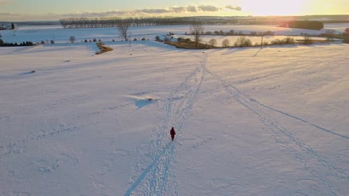 A man in a winter coat walks on a snowy road to a golden sunset with romantic clouds - aerial shot