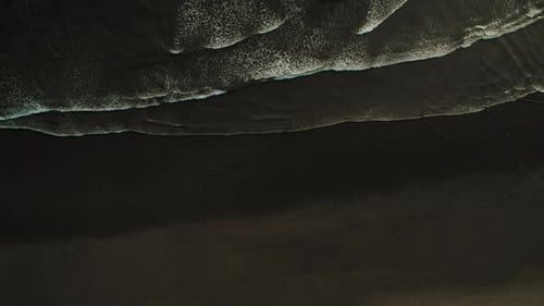 Aerial top view of black sandy beach with foamy waves forming patterns at shore