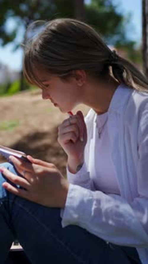 Young Woman Writing Notes in Notebook While Sitting in Peaceful Park Enjoying Nature's Tranquil