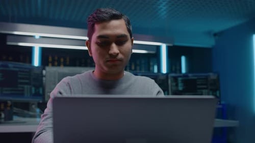 Portrait of Software Developer / Hacker / Programmer Sitting at His Desk and Working on Laptop Comp