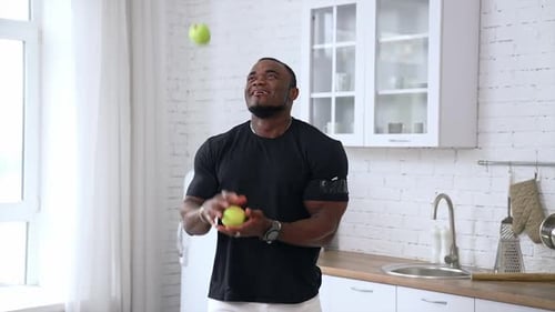 Happy young multiracial man joggling apples in kitchen background, selective focus, closeup.