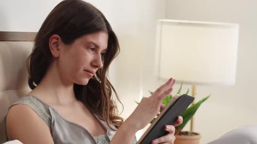 A Young Brunette Uses a Digital Tablet While Sitting on a Bed By the Window A Beautiful Longhaired