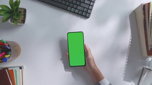 In der Ansicht von oben nach unten befindet sich ein Smartphone mit grünem Bildschirm auf einem weißen Schreibtisch