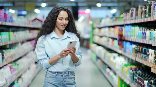 Happy young hispanic woman uses mobile phone in a supermarket while standing between the rows.