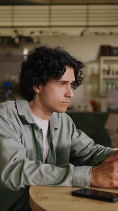Focused Young Man with Curly Hair Working Intently on a Laptop at a Wooden Table in a Cozy Modern