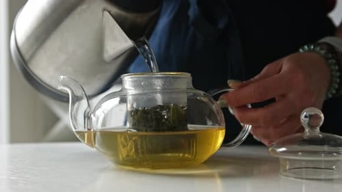 Girl Pouring Hot Water From The Kettle Into Teapot With Green Tea In The Kitchen