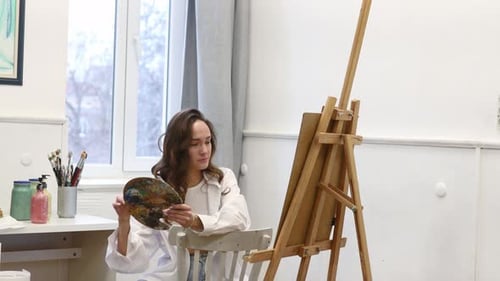 Young Female Artist Standing Near Easel and Holding Pencil While Working on Painting in Art Studio