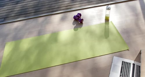 High angle view of yoga mat, laptop, bottle with juice and weights on floor at home, slow motion
