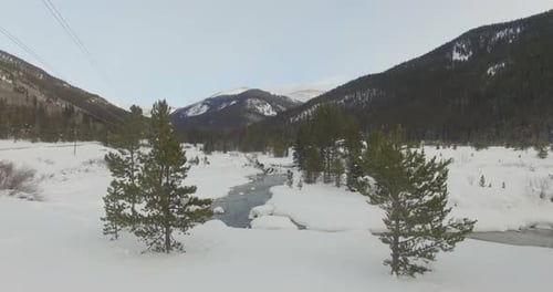 Flying over winding river in snow covered rocky mountain valley.