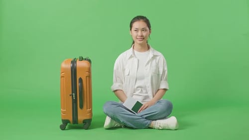 Full Body Of Asian Female Traveler With Luggage Looking Around While Sitting In The Green Screen