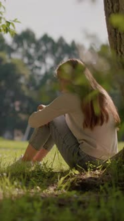 Reflective Woman Sitting on Grass in Sunlit Park with Tranquil Background