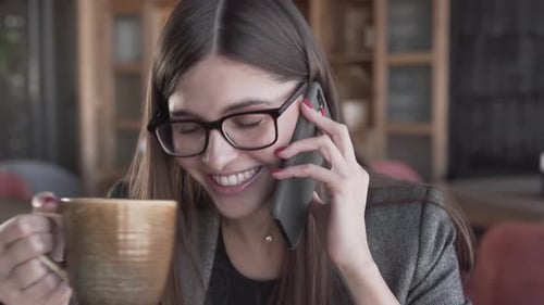 Beautiful young woman in a glasses is sitting in a cafe, drinking a coffee and talking on a phone. W