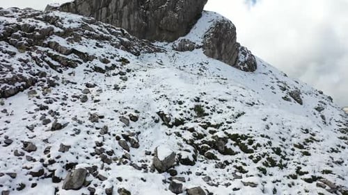 Aerial shot flying upwards revealing a massive cliff on top of the snowy mountain with a man standin