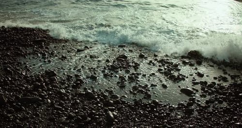 Black pebble beach with muddy ocean waves crashing on rocky shore creating foam in slow-motion. Stor