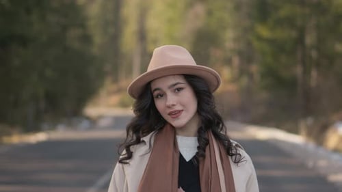 Stylish Young Woman in Beige Hat and Scarf Standing Outdoors on a Forest Road Looking Down with a