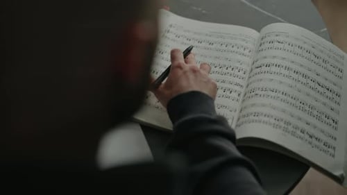 Back Top View of Music Composer Writing Melodic Notes Sitting at Table with Laptop and Sheet Music